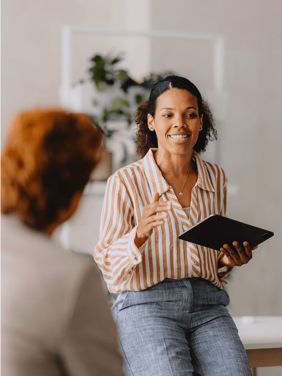 Frau in gestreifter Bluse spricht mit Kollegin und hält Tablet in der Hand, Bürohintergrund mit Pflanzen.