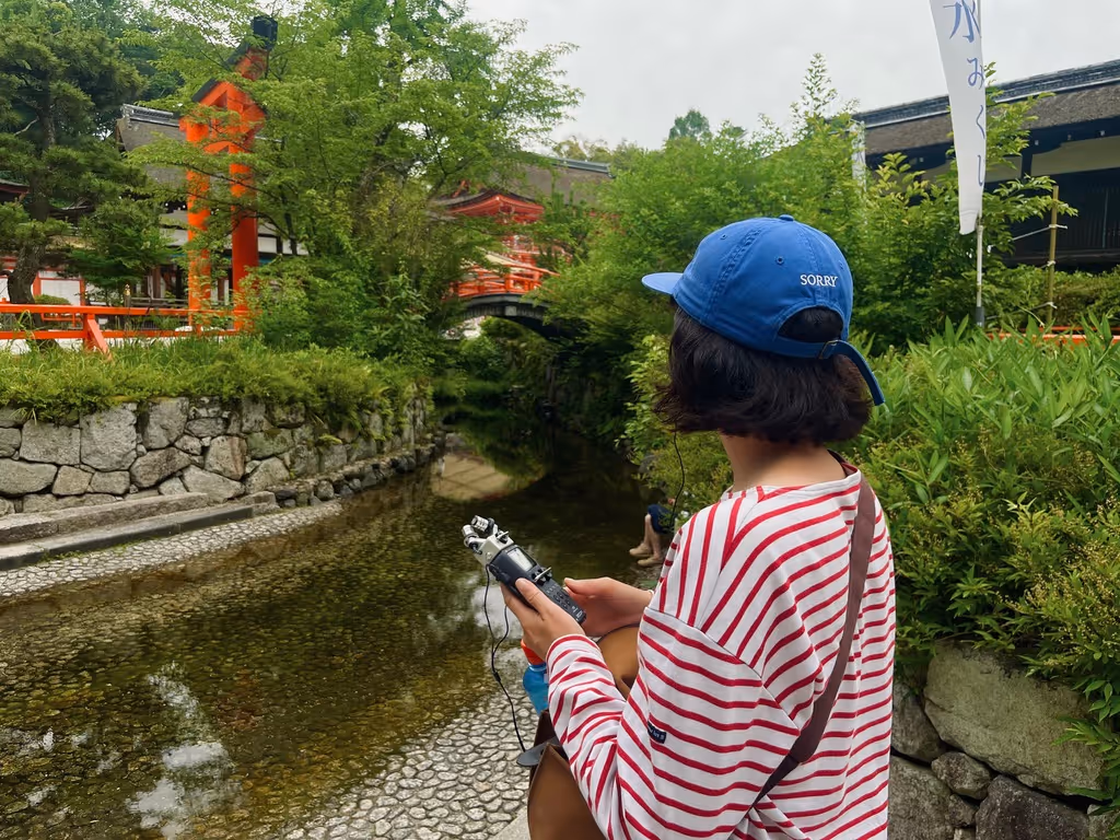 A photograph of a recent sound walk guest who joined a Kyoto, in Sound walk. This photograph is from Shimogamo Shrine in Kyoto and features their famous bridge that runs over a river. A young lady stands in the foreground holding a sound recorder.