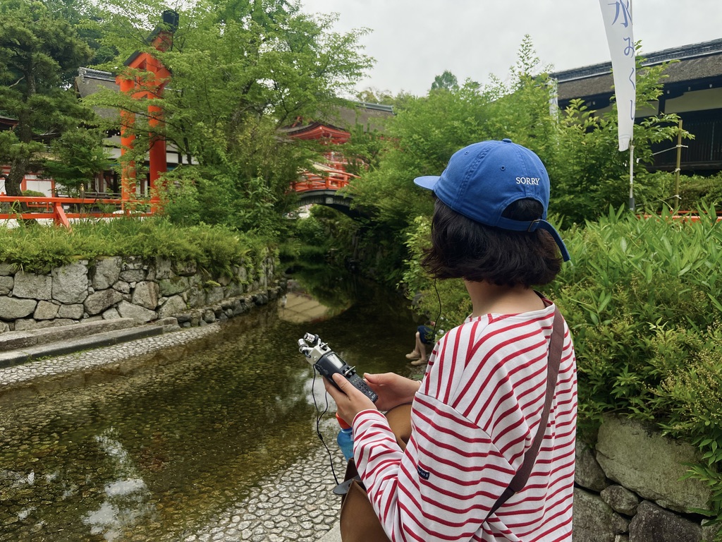 A photograph of a recent sound walk guest who joined a Kyoto, in Sound walk. This photograph is from Shimogamo Shrine in Kyoto and features their famous bridge that runs over a river. A young lady stands in the foreground holding a sound recorder.