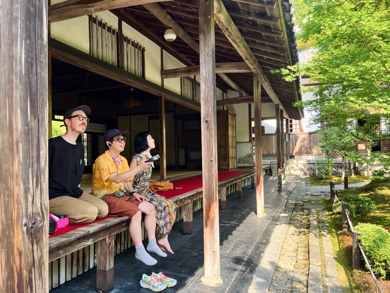 A photograph of a recent sound walk guest who joined a Kyoto, in Sound walk. This photograph features three people looking out from the veranda of a temple in Kyoto into its garden on the right of the picture. One of them is holding a sound recorder.