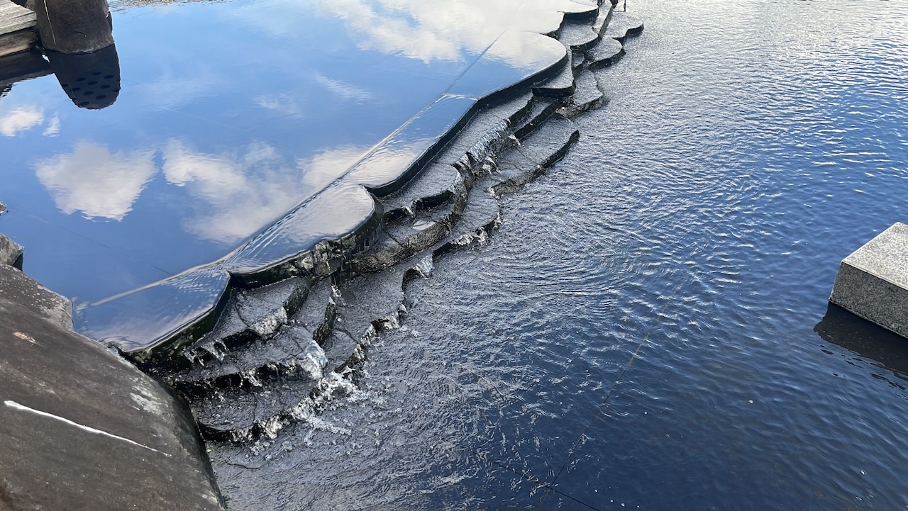 A photograph of the Mirror Pool in the Suzaku Garden in Kyoto. Clouds from a blue sky above are beautifully reflected in the black surface of the water which is running down a slight waterfall.