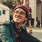 A man with glasses and a beanie smiles while riding a bicycle outdoors.
