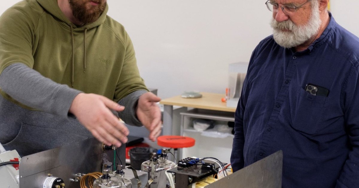 A man gestures toward a small machine that's in creation. Another man in a blue button-down watches him.