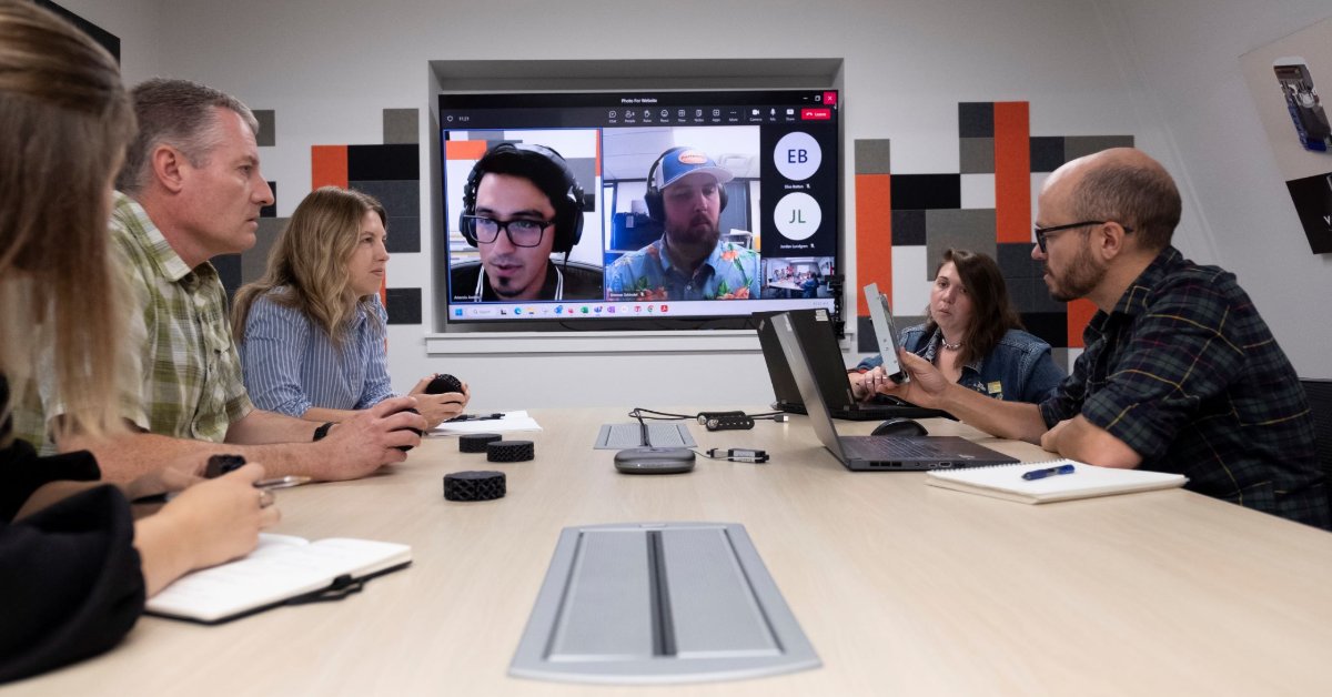 A group of people sit around a table and chat, while other folks look on from an on-screen Microsoft Teams call.