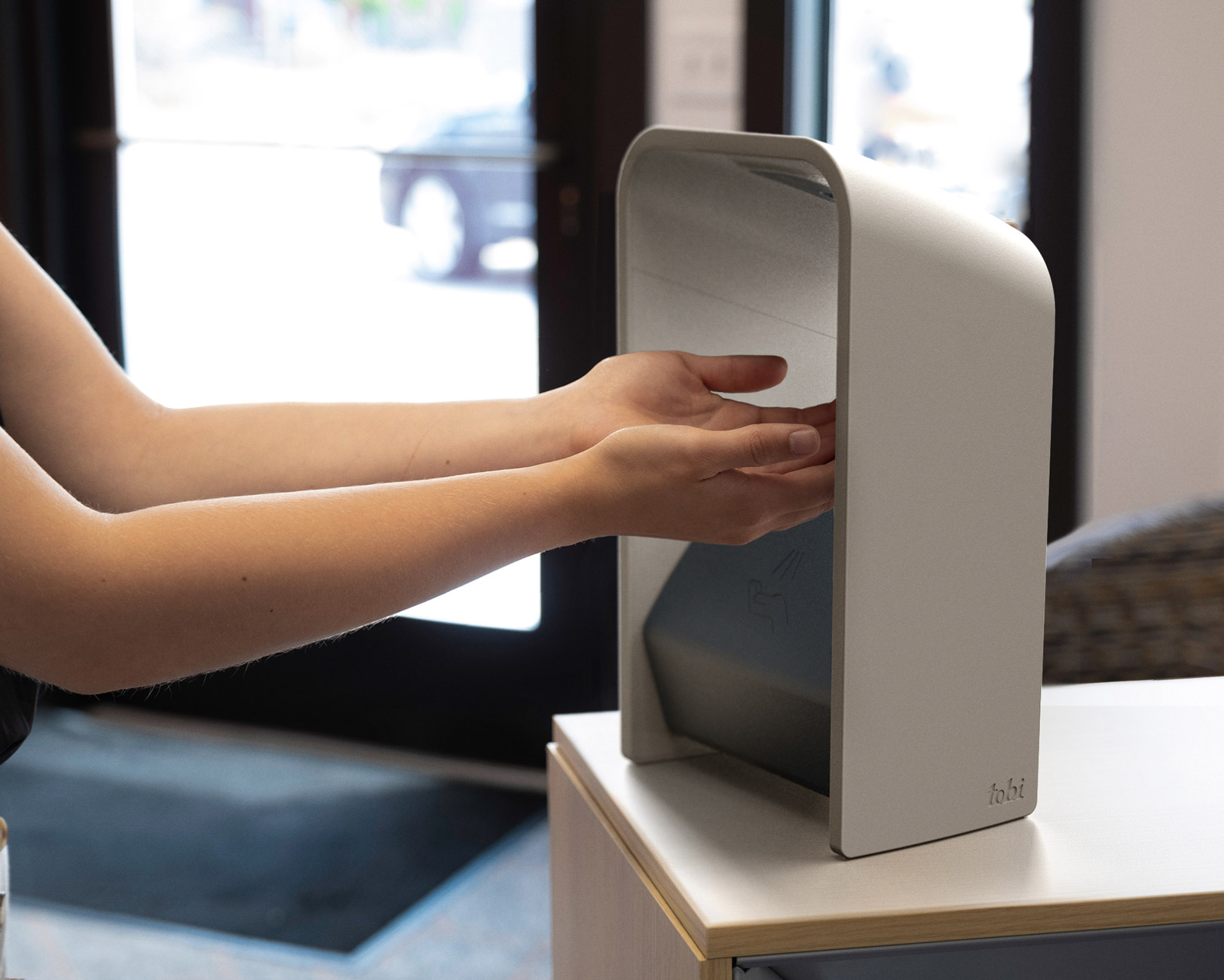 Person places both hands inside a touch-free hand scanner on a countertop near a glass entrance and reception area.
