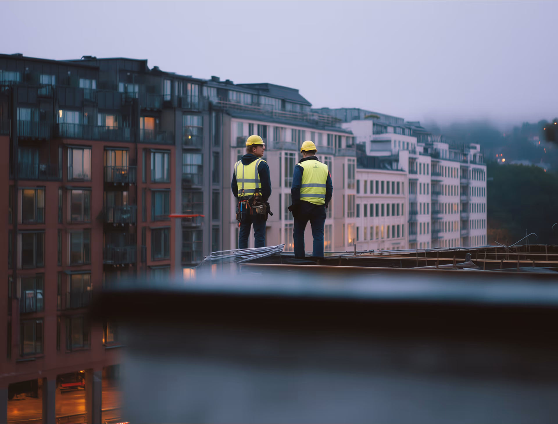 To bygningsarbeidere i refleksvester og hjelmer står på et tak med utsikt over et bybilde i den tidlige morgentåken.