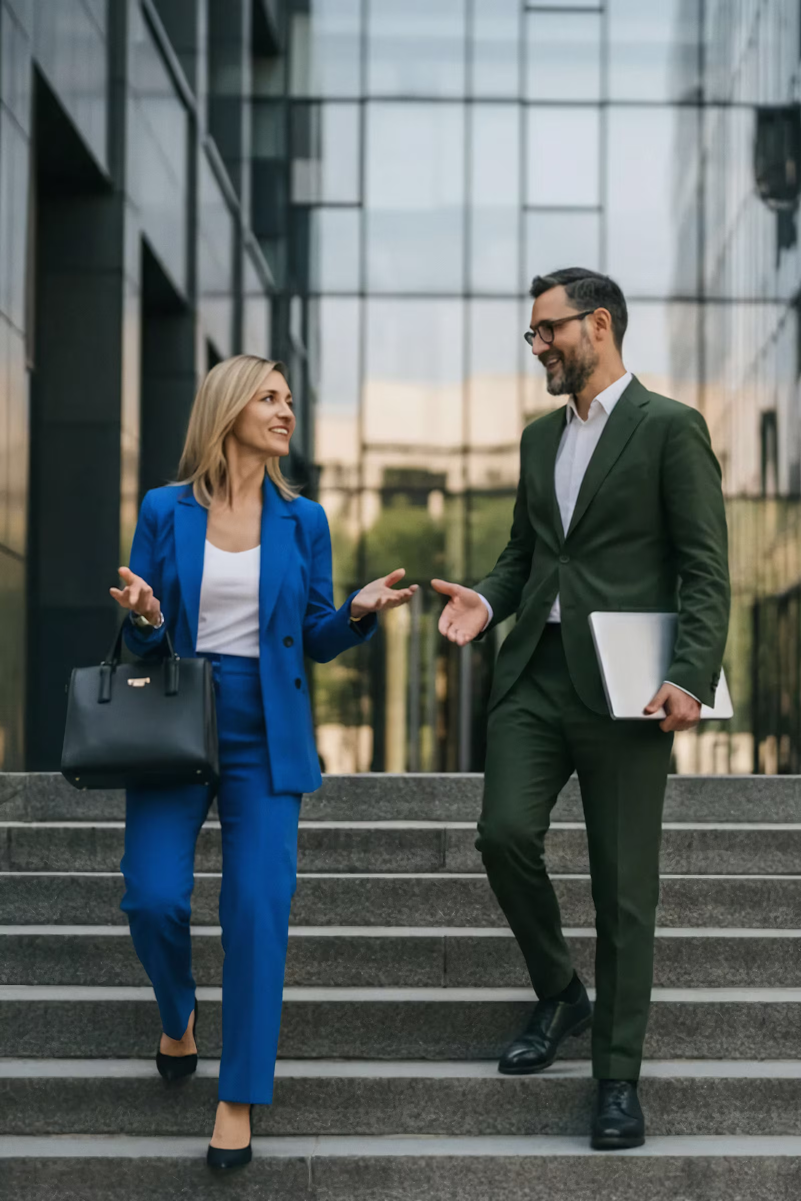 Eine Frau im blauen Anzug und ein Mann im grünen Anzug unterhalten sich lachend beim Hinabgehen von Außentreppen vor einem modernen Glasgebäude.