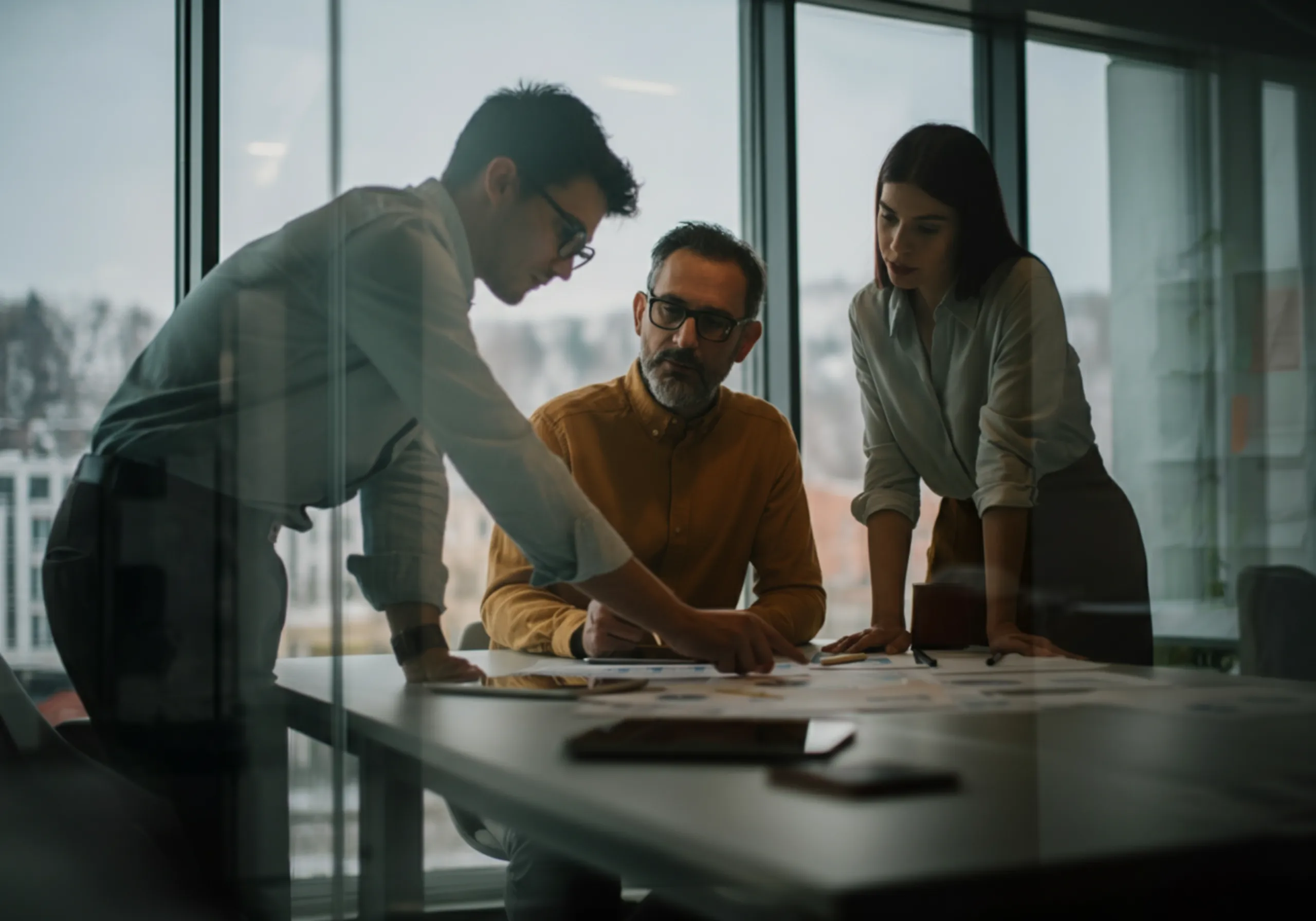 Drei Personen in einem modernen Büro besprechen Unterlagen auf einem Tisch vor großen Fenstern.