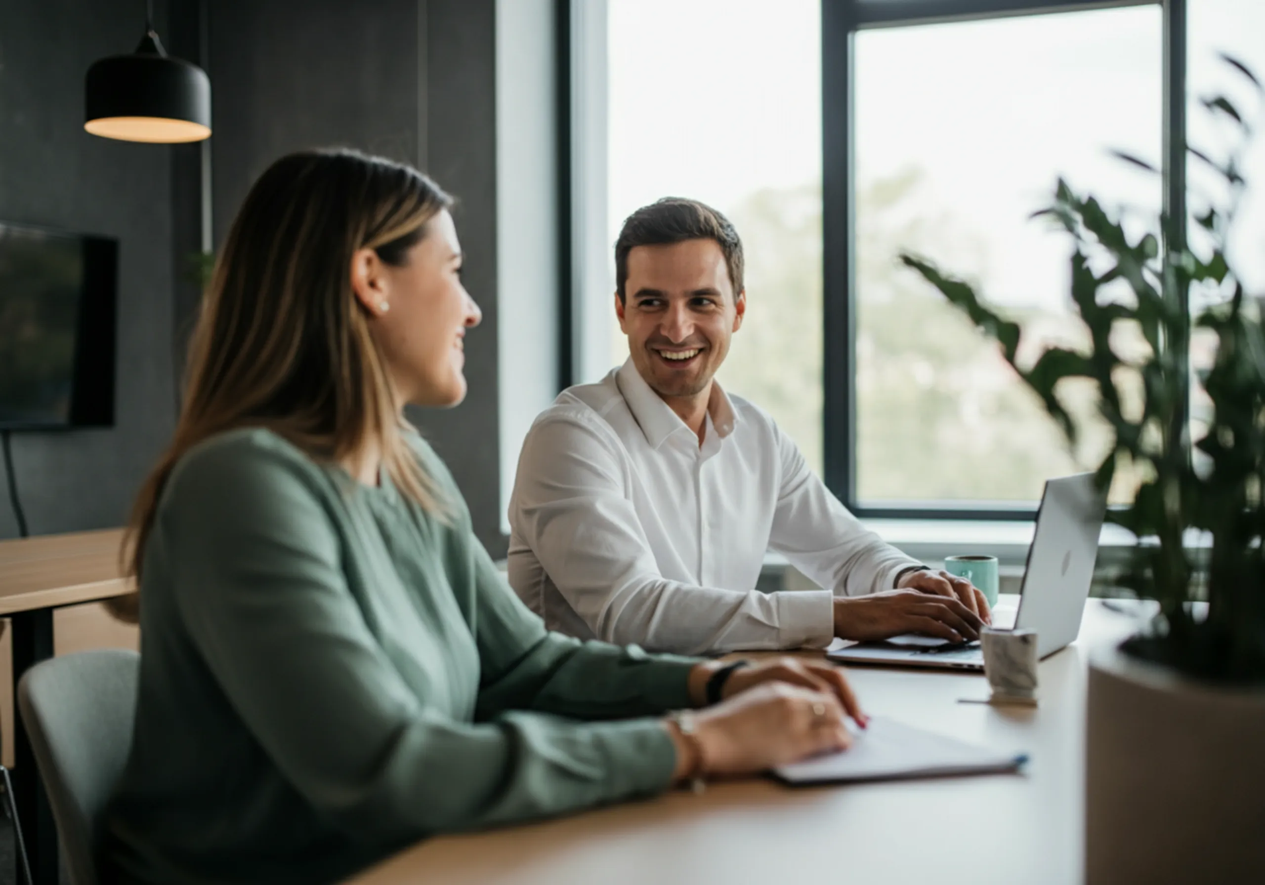 Ein Mann und eine Frau sitzen an einem Tisch in einem modernen Büro und unterhalten sich dabei lächelnd, der Mann hat einen Laptop vor sich.