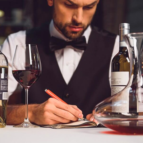 Sommelier writing notes at a table with wine bottles, a decanter, and a glass of red wine.