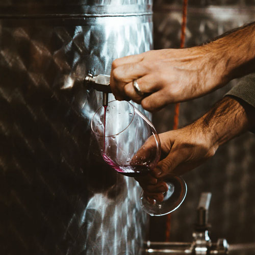 Person pouring red wine from a stainless steel tank into a glass.