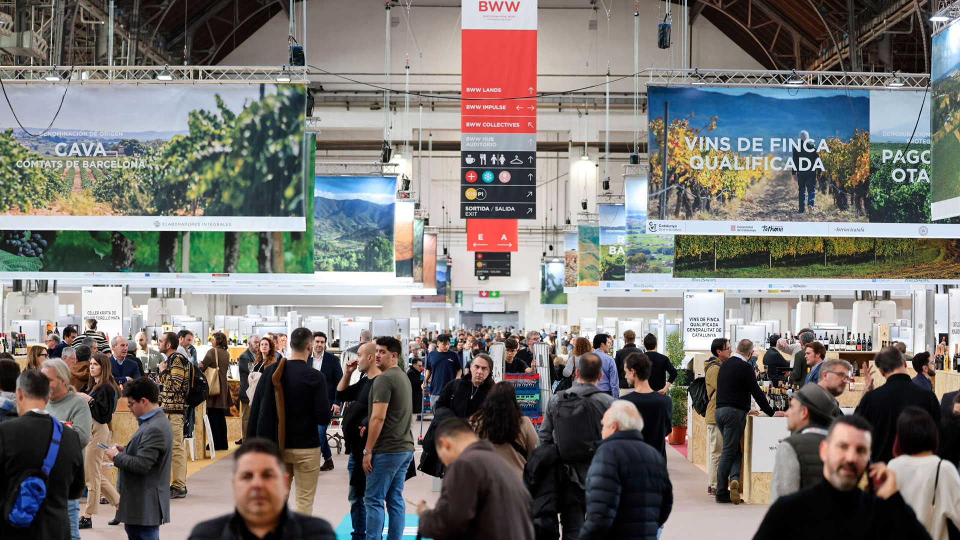 Interior view of Barcelona Wine Week showing a busy central aisle with wine professionals, exhibitor booths, and large hanging banners highlighting Spanish wine appellations.
