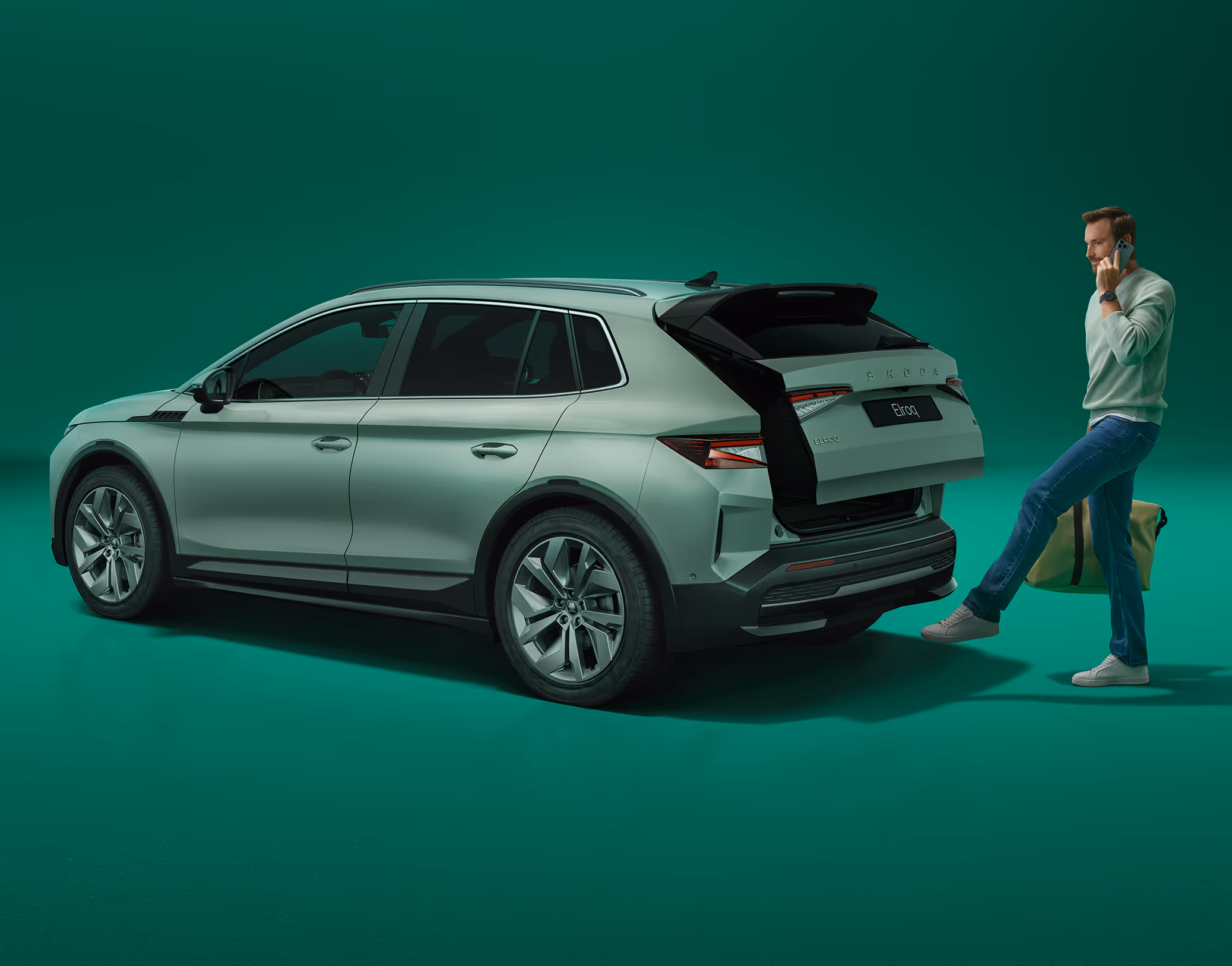 Ein Mann öffnete berührungslos den Kofferraum eines Škoda Elroq in der Farbe Timiano-Grün, indem er einen Fuß unter den Sensor hält. Die Person telefoniert mit einer Hand, in der anderen trägt sie eine Tasche.