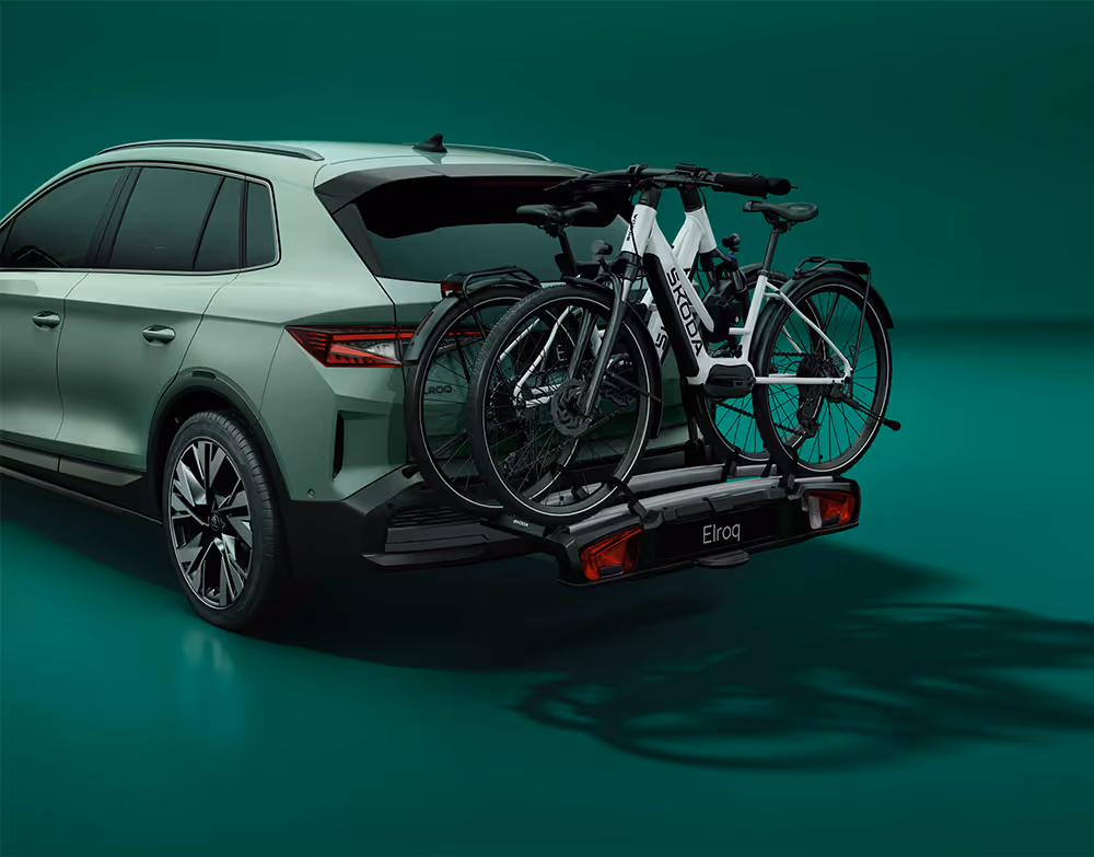 Heckansicht eines Škoda Elroq mit einem Fahrradträger auf der Anhängerkupplung vor grünem Studiohintergrund. Auf dem Träger sind zwei Fahrräder mit Škoda Schriftzug auf dem Rahmen montiert.