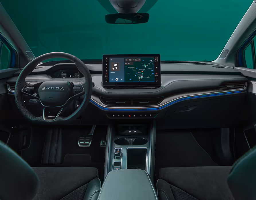 Blick über die Mittelarmlehne auf das Lenkrad, das Infotainmentsystem und das Armaturenbrett im Škoda Elroq Sportline. Blaue Ambient-Beleuchtung.
