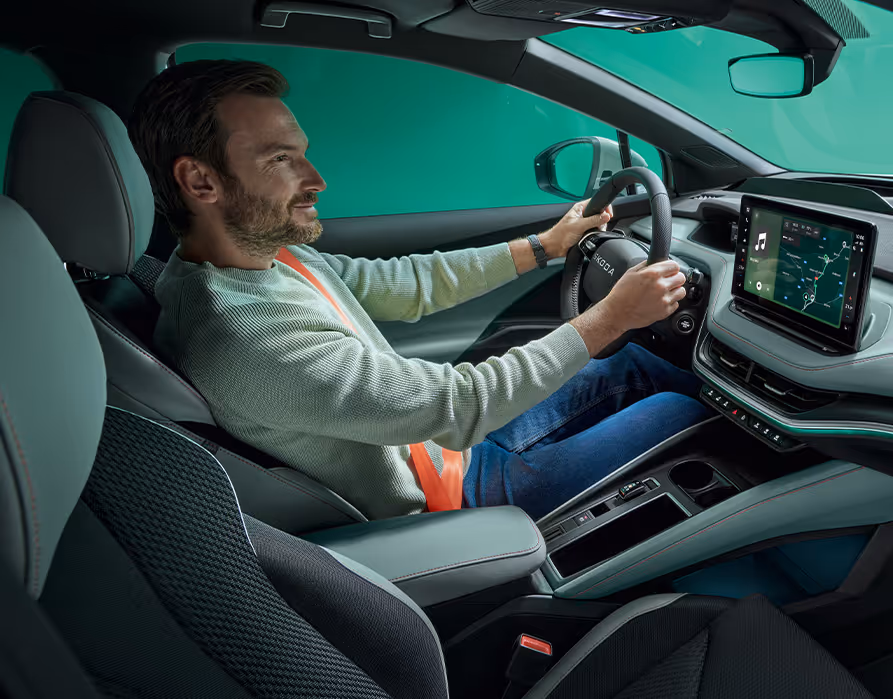 Seitlicher Blick in den vorderen Innenraum des Škoda Elroq, in dem ein Mann am Steuer sitzt. Ein markantes Detail ist der leuchtend orangefarbene Sicherheitsgurt, der einen starken farblichen Kontrast zu den ansonsten dunkel gehaltenen Sitzen und der Mittelkonsole bildet. Das moderne Cockpit wird durch ein großes, freistehendes Infotainment-Display und das Multifunktionslenkrad mit dem neuen Škoda Schriftzug geprägt.