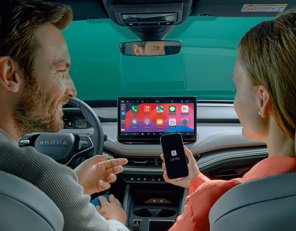 Ein Mann im Fahrersitz interagiert mit der Person auf dem Beifahrersitz.  Auf dem Infotainment-Display sind die Icons verschiedener Smartphone-Apps zu sehen, was die SmartLink-Konnektivität ausdrücken soll.
