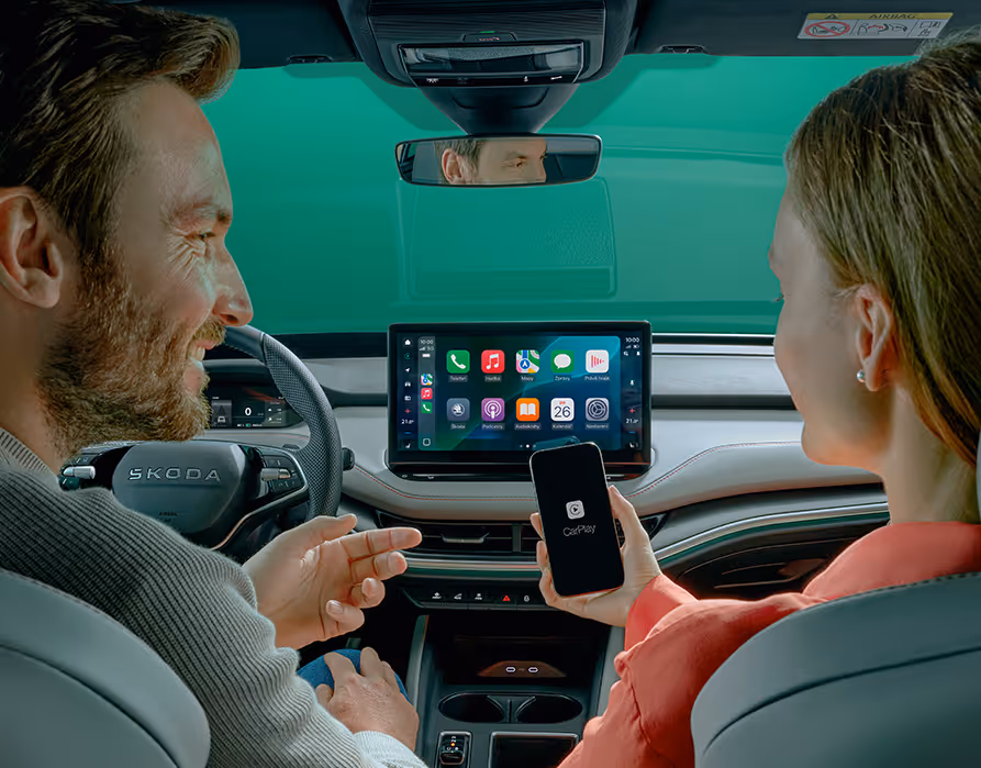 Blick zwischen den Vordersitzen in das Cockpit des Škoda Elroq, in dem Fahrer und Beifahrerin lächelnd auf das zentrale Infotainment-Display blicken. Die Beifahrerin hält ein Smartphone in der Hand, welches über Apple CarPlay mit dem Fahrzeug verbunden ist und dessen App-Oberfläche auf dem großen Touchscreen gespiegelt wird. Diese Darstellung veranschaulicht die nahtlose Integration des Smartphones in das digitale Bediensystem des Fahrzeugs.