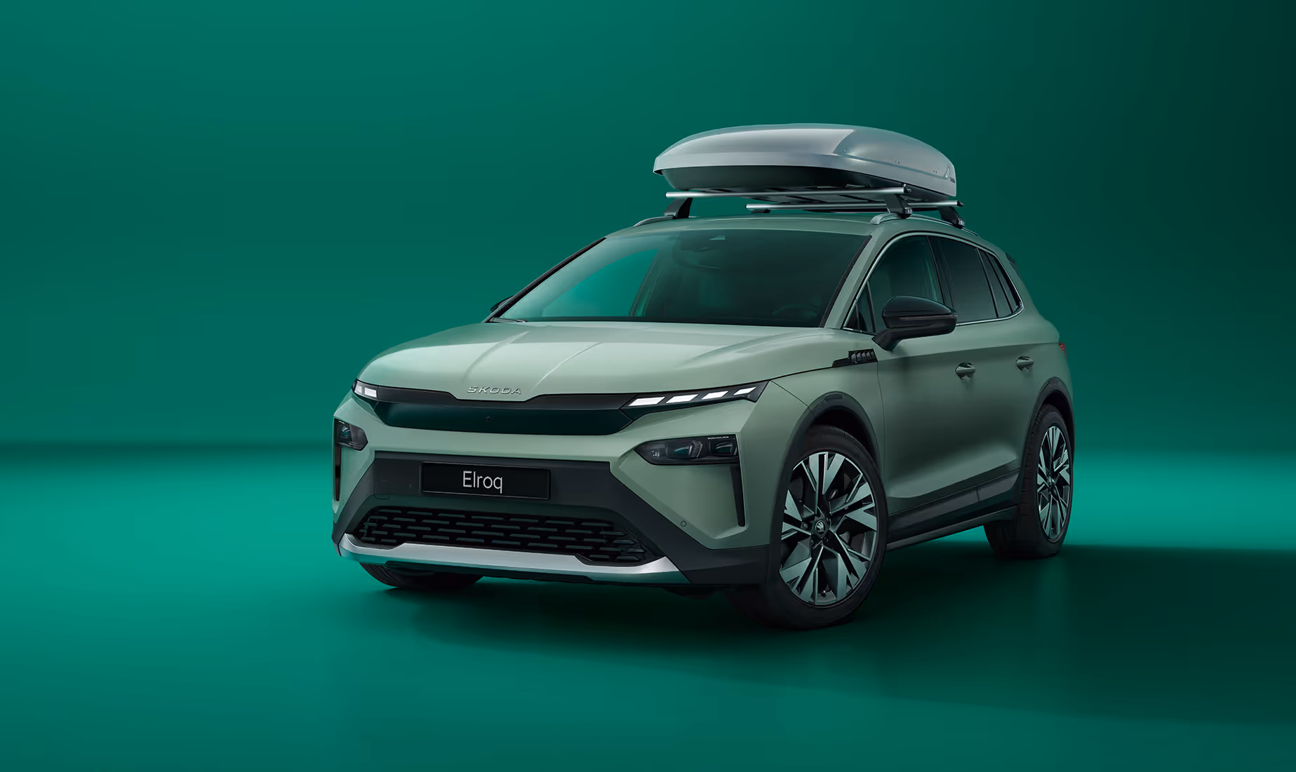 Frontalansicht eines Škoda Elroq mit einer Dachbox vor grünem Studiohintergrund. Das Fahrzeug ist für eine Reise mit zusätzlichem Gepäck ausgestattet.
