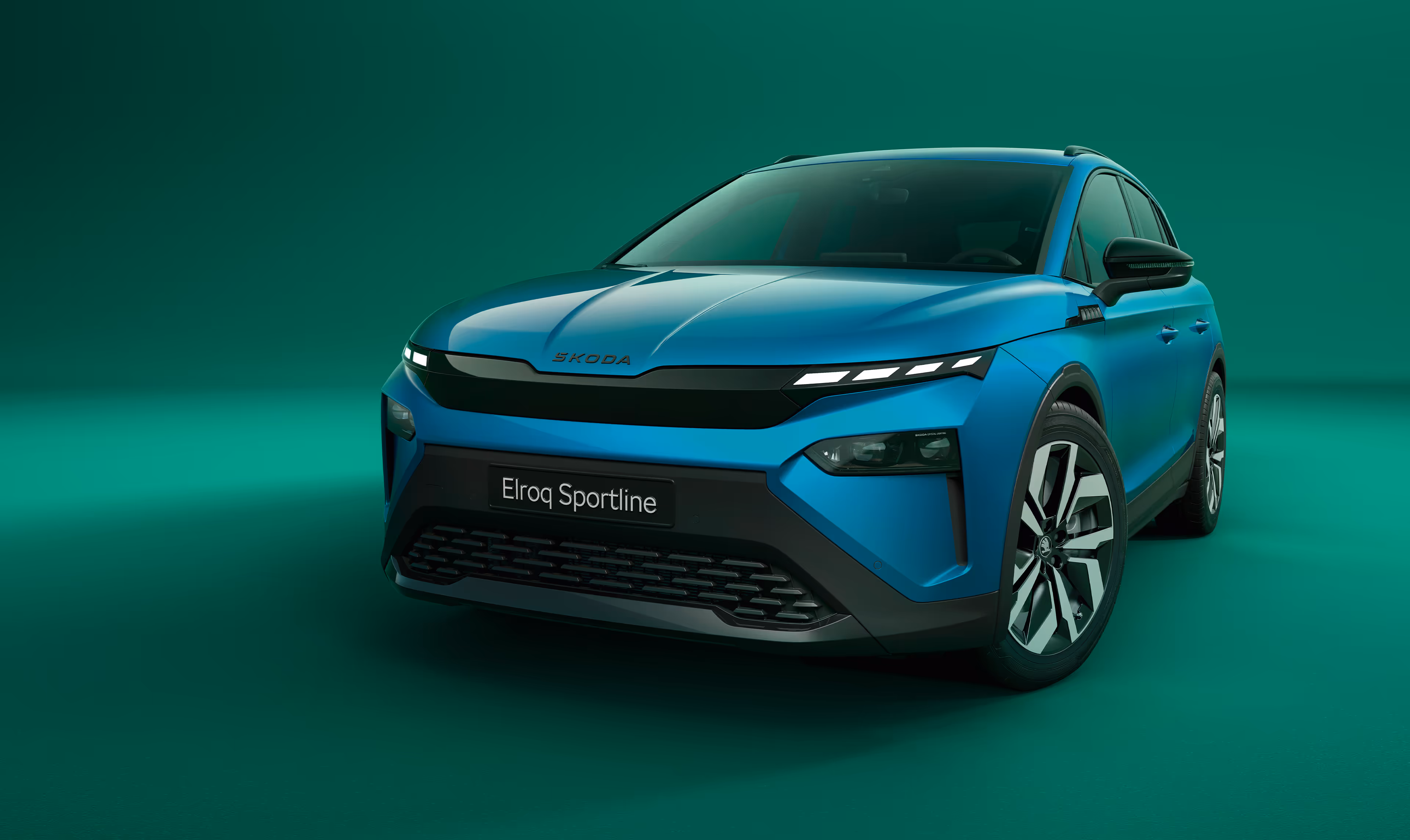 Frontalansicht des Škoda Elroq Sportline in der Farbe Race-Blau Metallic vor grünem Studiohintergrund. Das Fahrzeug zeichnet sich durch schwarze Designelemente am Kühlergrill und an der Frontschürze aus.