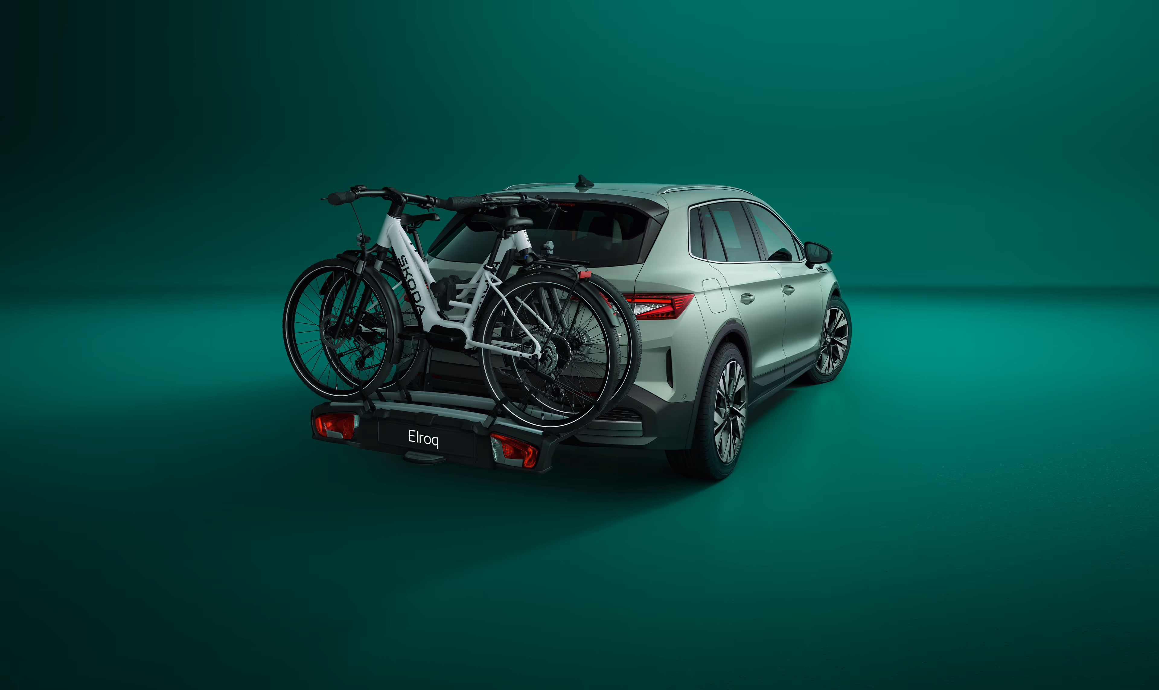 Heckansicht eines Škoda Elroq mit einem Fahrradträger auf der Anhängerkupplung vor grünem Studiohintergrund. Auf dem Träger sind zwei Fahrräder mit Škoda Schriftzug auf dem Rahmen montiert.