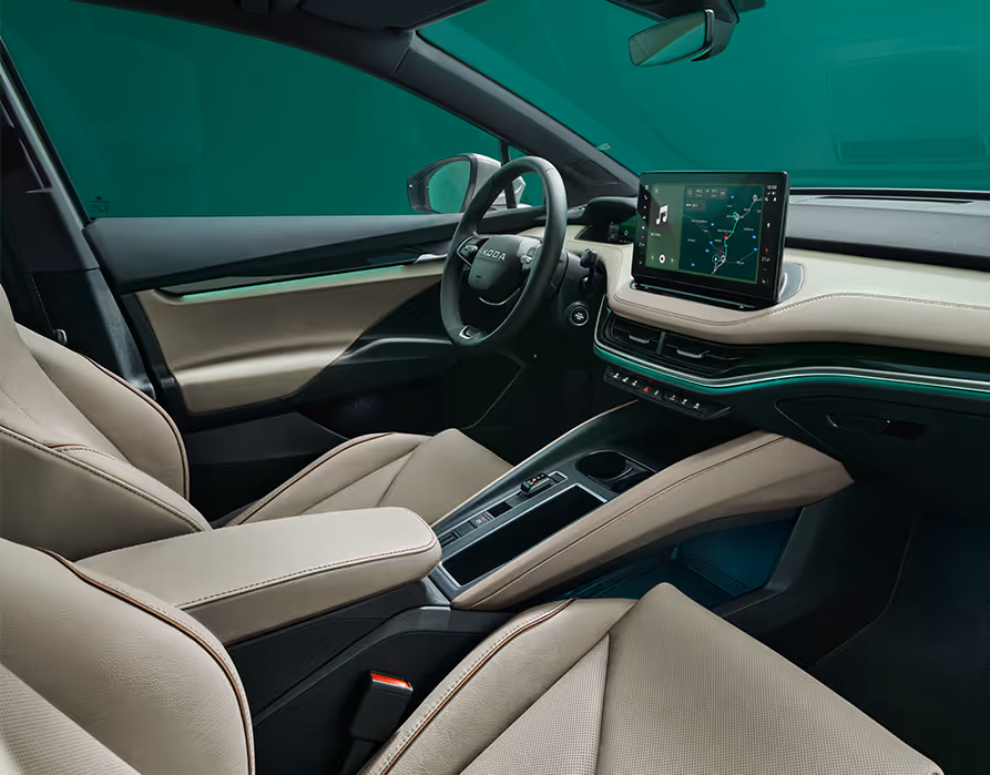 Ein Blick in den vorderen Innenraum des Škoda Elroq mit Fokus auf den Fahrer- und Beifahrersitz. Die Sitze der Design Selection „L&K“ sind mit beigem Leder bezogen und weisen cognacfarbene Ziernähte auf. Die grüne Ambientebeleuchtung leuchtet entlang des Armaturenbretts und der Fahrertür.