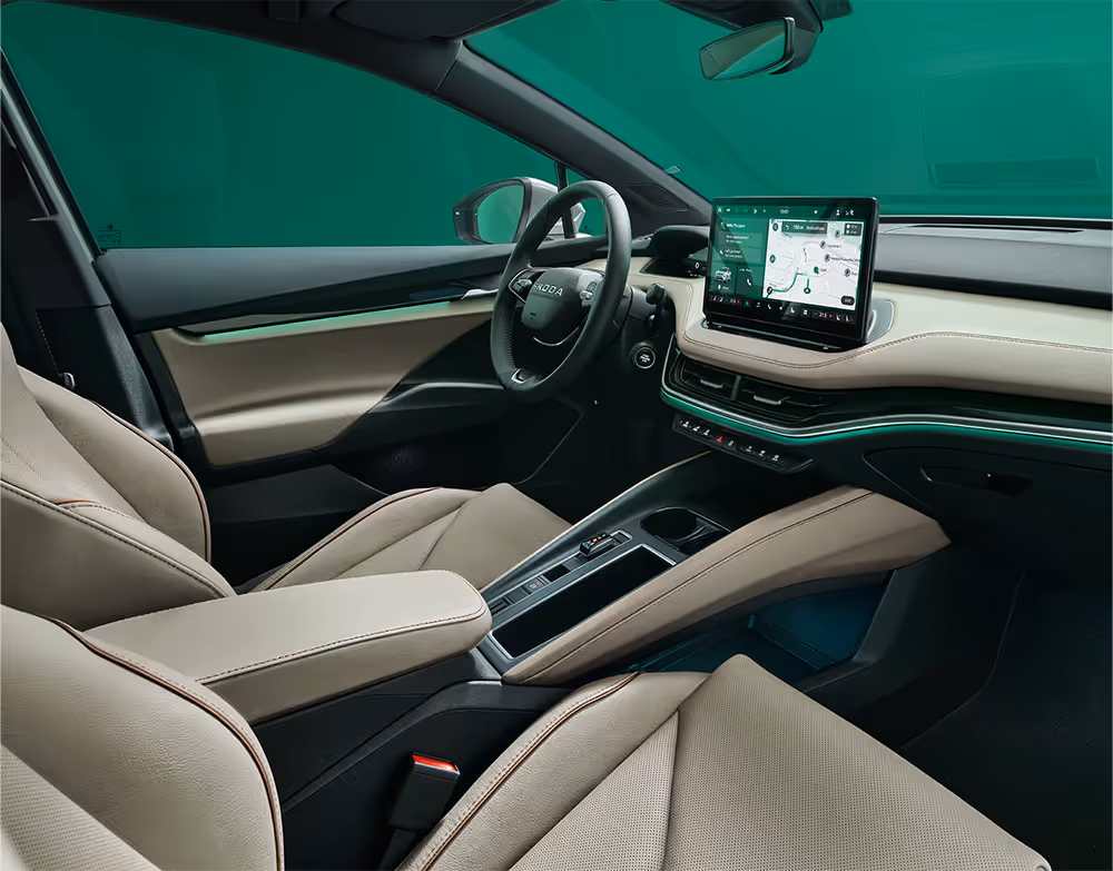 Ein Blick in den vorderen Innenraum des Škoda Elroq mit Fokus auf den Fahrer- und Beifahrersitz. Die Sitze der Design Selection „L&K“ sind mit beigem Leder bezogen und weisen cognacfarbene Ziernähte auf. Die grüne Ambientebeleuchtung leuchtet entlang des Armaturenbretts und der Fahrertür.