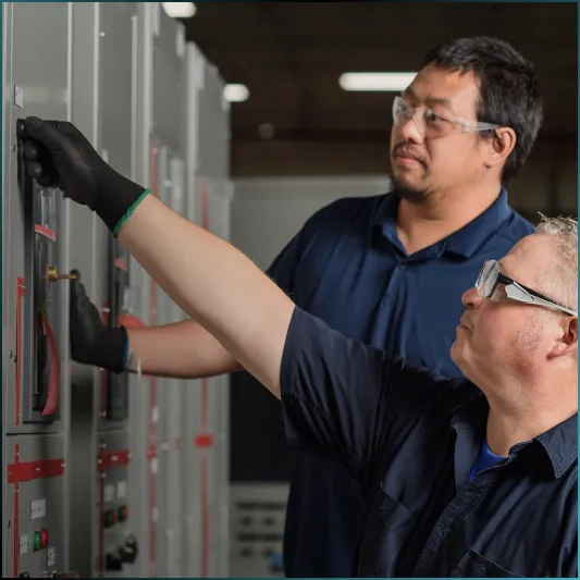 Two men working on electrical control panels wearing gloves and safety glasses in a technical setting.
