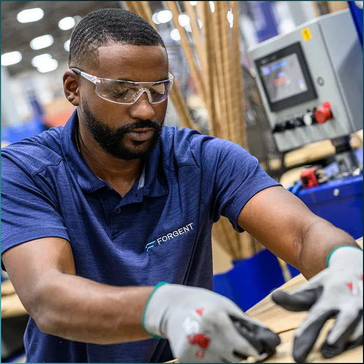 Man in safety glasses and Forgent shirt working with hands on a wooden surface in a workshop.