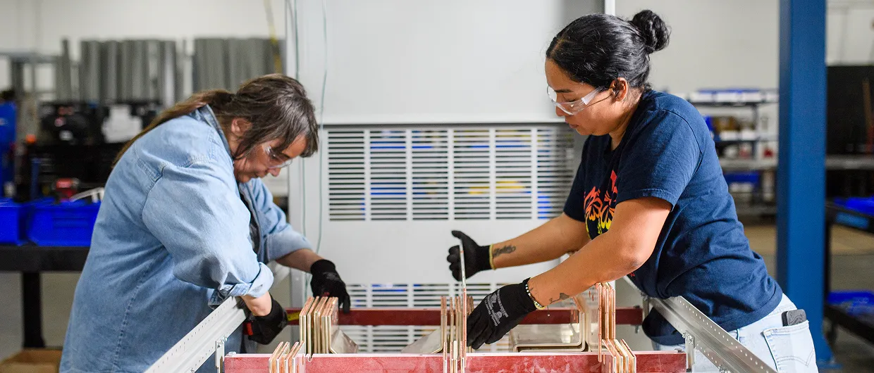 Two women wearing gloves and safety glasses working with equipment in an industrial setting.