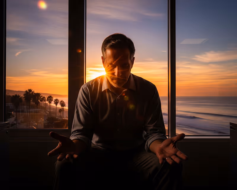 A focused man sitting in front of a window with his hands clasped.