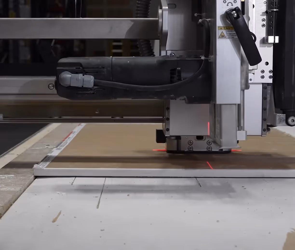 Close-up of an automated cutting machine with red laser guides cutting a large sheet of drywall on a flat surface.