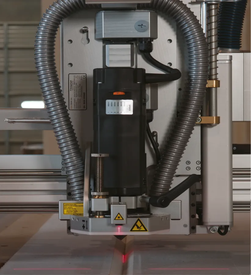 Close-up of a CNC machine spindle with protective hoses and laser alignment lights over a piece of wood being cut.