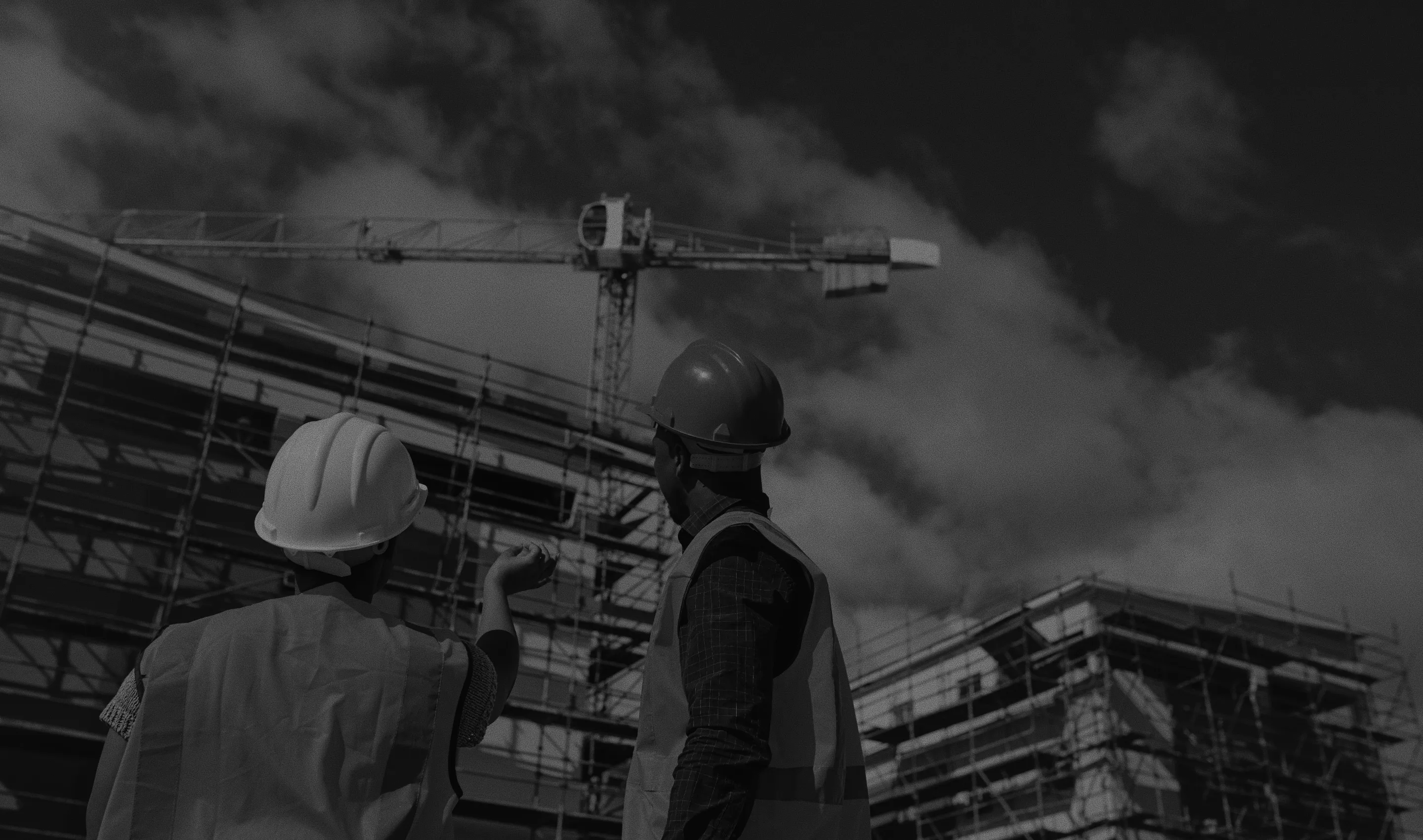 Two construction workers wearing hard hats and reflective vests discussing at a building site with scaffolding and a crane in the background.