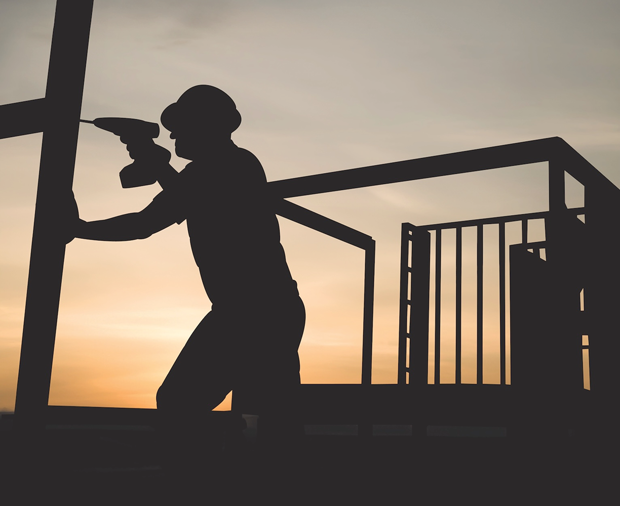 Silhouette of a construction worker using a power drill on a metal frame during sunset.