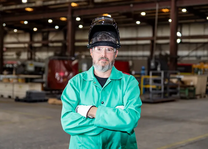 Factory worker in safety gear and a green jacket standing with arms crossed inside an industrial facility