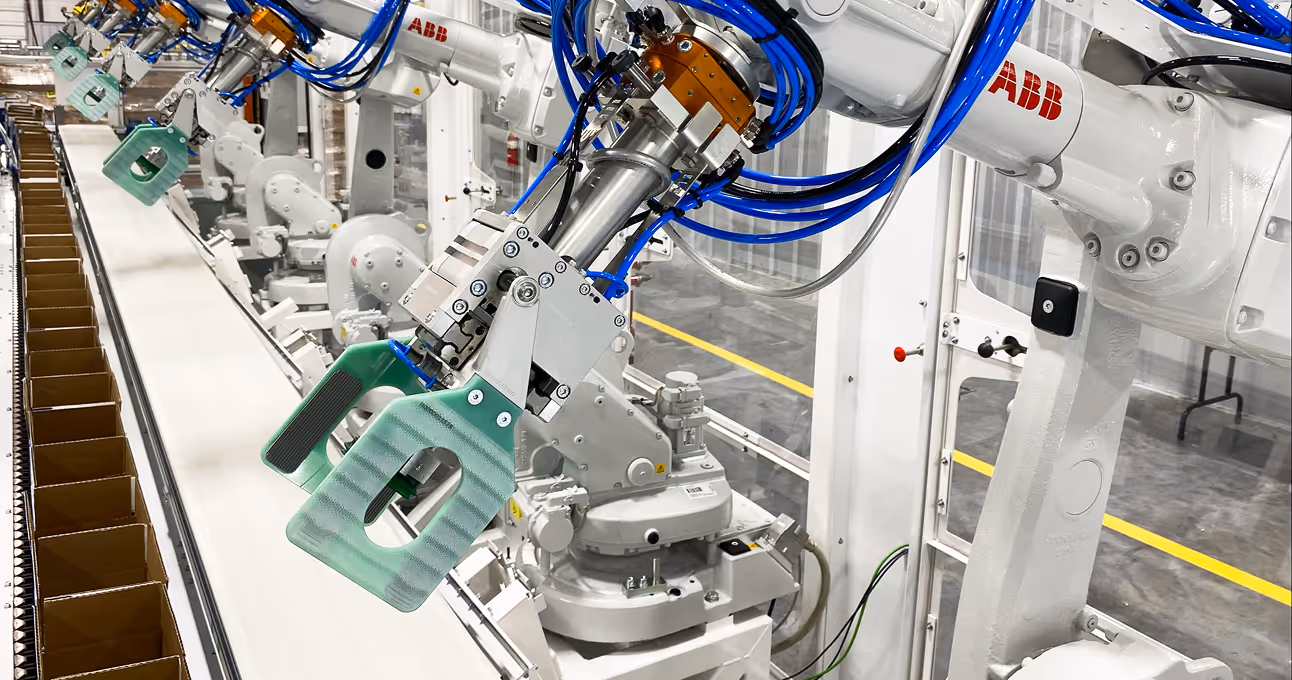 Row of white ABB industrial robotic arms with blue cables, each equipped with green grippers, assembling or packaging along a conveyor belt with empty brown boxes.