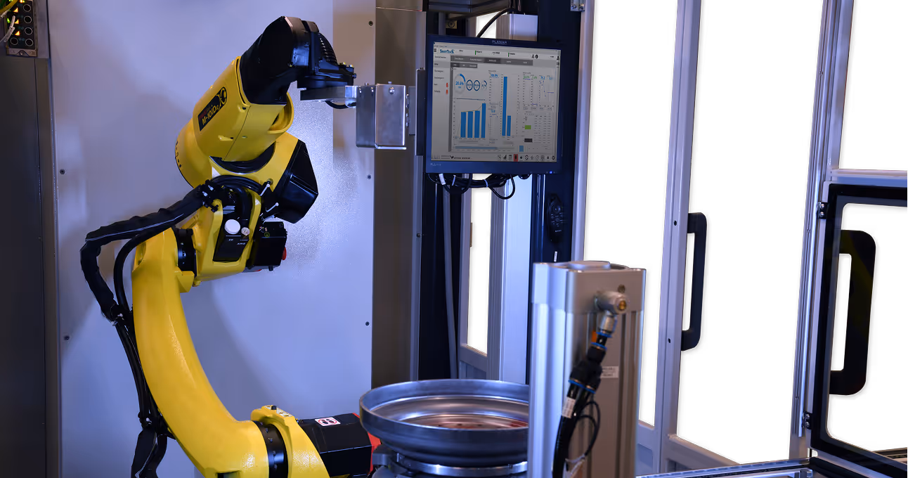 Yellow robotic arm operating in an industrial setting with a monitor displaying data in the background.