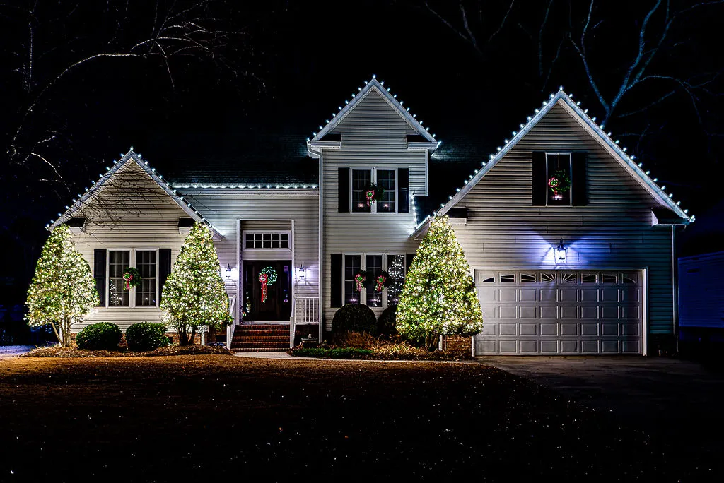 A white two-story house in Greenville, NC decorated by Christmas light installers in Greenville, NC glowing white Christmas lights outlining the roof and windows, with wreaths on the front door and windows, and bushes and trees in front wrapped in bright white lights, illuminated at night.