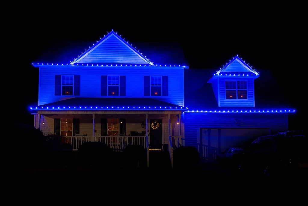 Two-story house in Greenville, NC decorated with blue Christmas lights outlining the roof and dormer windows at night, with a lit wreath on the front door.