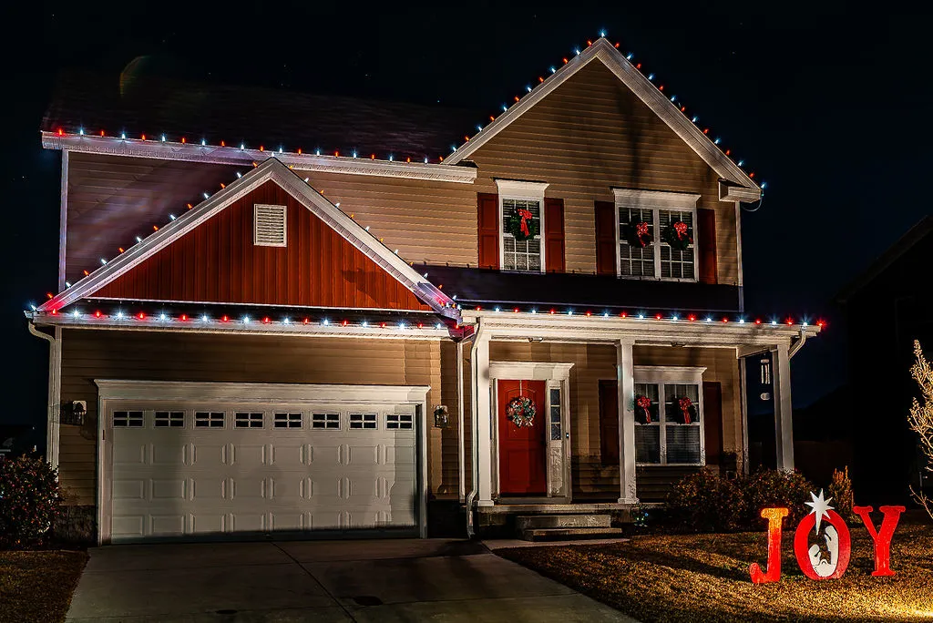 Two-story Greenville, NC house at night decorated with red, white, and blue Christmas lights installed by Walston Evans along the roof edges, red bows and greenery wreaths on six windows, a wreath on the red front door, and illuminated red letters spelling JOY in the front yard.