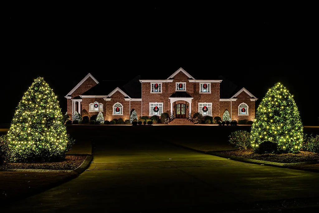 A large brick house in Greenville, NC with Christmas lights installed at night, with six wreaths on the windows and door, and two large shrubs on either side of the driveway covered in white Christmas lights.