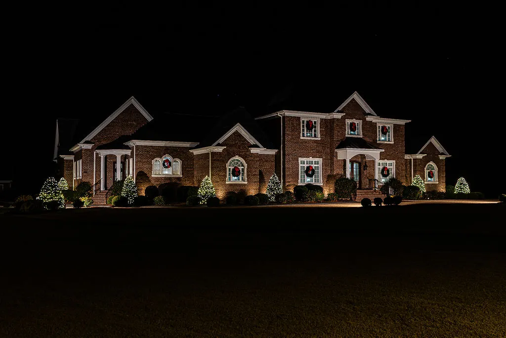 A large brick house in Greenville, NC at night decorated with Christmas lights and wreaths on every window and bushes and small trees adorned with white holiday lights.
