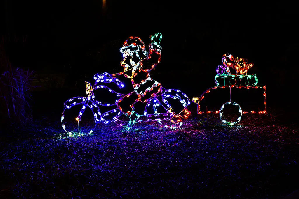 Christmas light decoration in Greenville, NC featuring Santa Claus riding a motorcycle with a trailer labeled 'TOYS' carrying candy canes and a teddy bear, all illuminated in multicolored lights against a dark background.