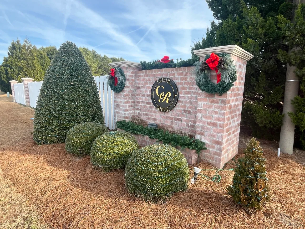 Brick entrance sign for Cedar Ridge with gold lettering on a black oval plaque, decorated with two Christmas wreaths with red bows and pine cones, surrounded by manicured bushes and pine straw covering the ground under a blue sky with wispy clouds.