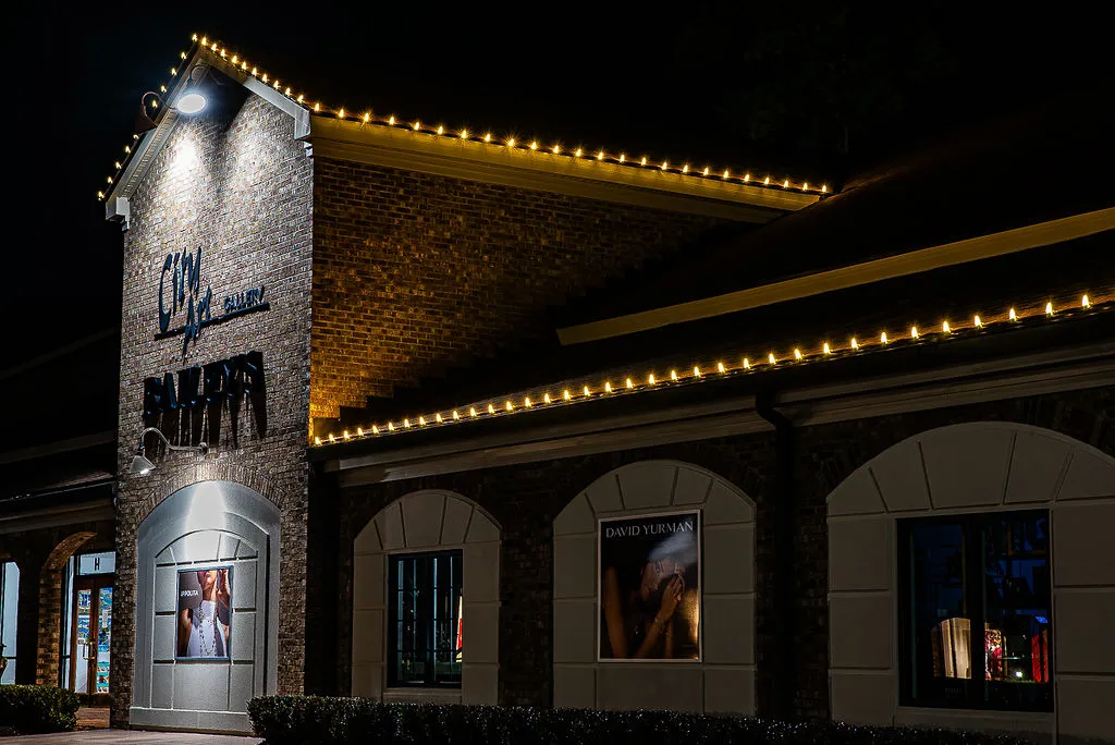Exterior of a brick art gallery building at night with warm string lights along the roof edges, illuminated signage reading 'City Art Gallery,' and posters visible through windows.