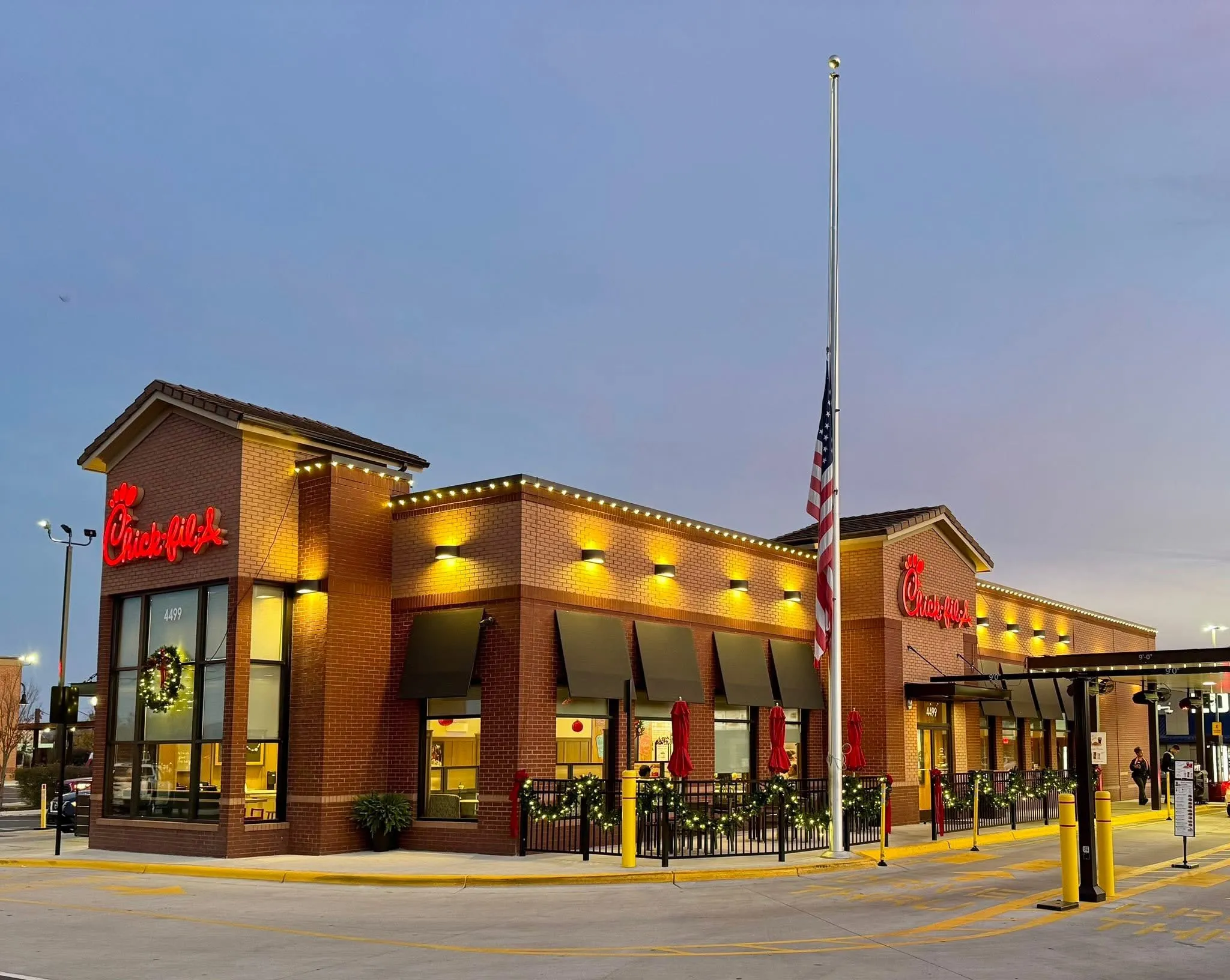 Chick-fil-A Kinston restaurant building at dusk with illuminated exterior lights, red signage, holiday wreath on window, festive garlands decorating the outdoor seating area all installed by Walston Evans Home Services, and an American flag flying at half-mast.