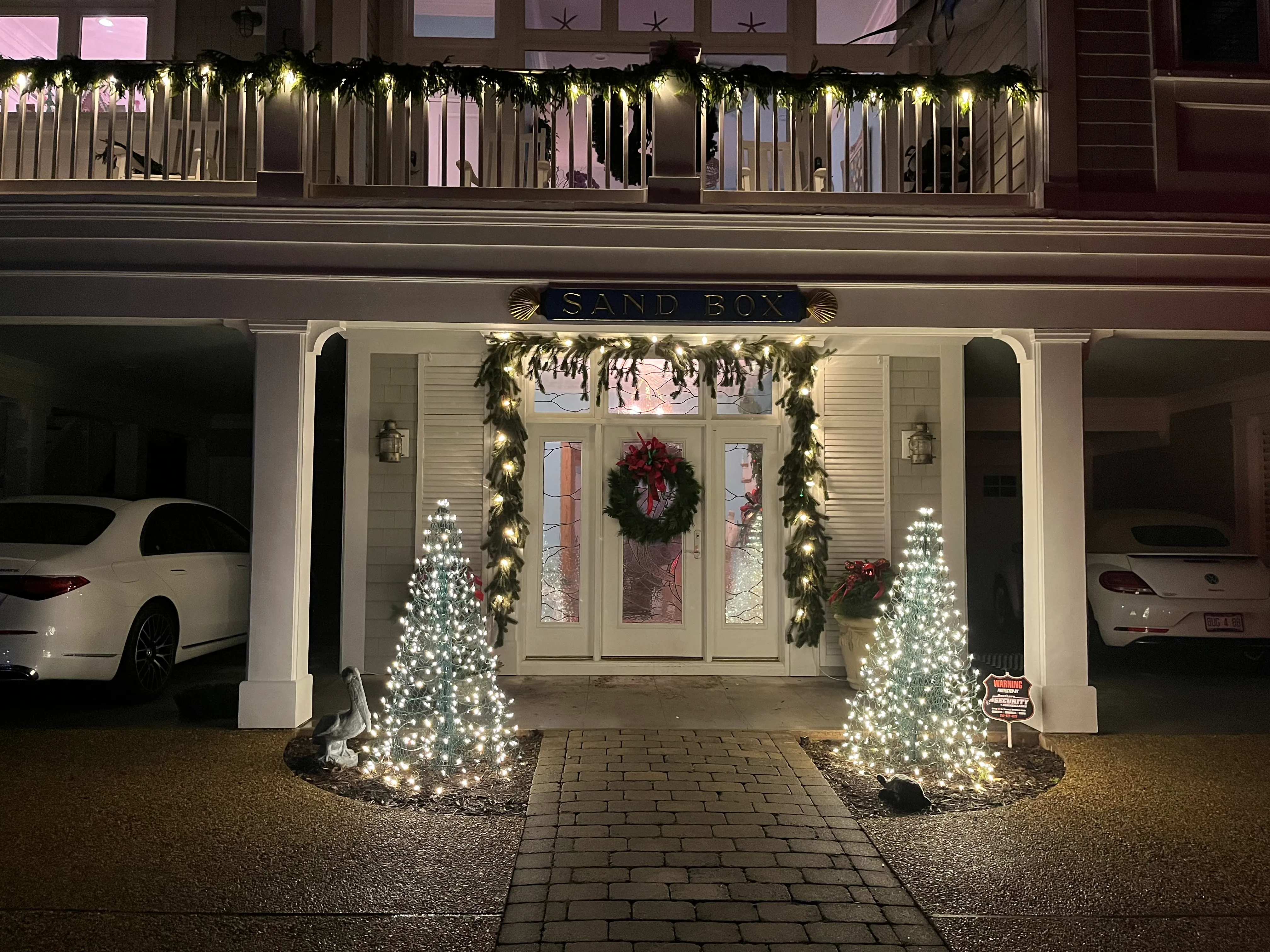 Front entrance of an Atlantic Beach house decorated with holiday lights and greenery, including two small lit Christmas trees beside the walkway, a wreath on the door, and garlands with lights around the door frame and balcony railing above. Two cars are parked under the house's covered area on each side.