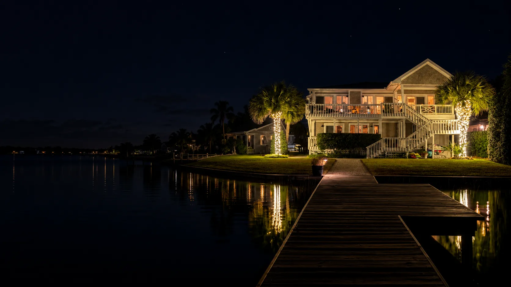 Night view of an Atlantic Beach waterfront house with warm lights glowing from windows and palm trees wrapped in string lights installed by Walston Evans Home Services, reflecting on calm water next to a wooden dock.