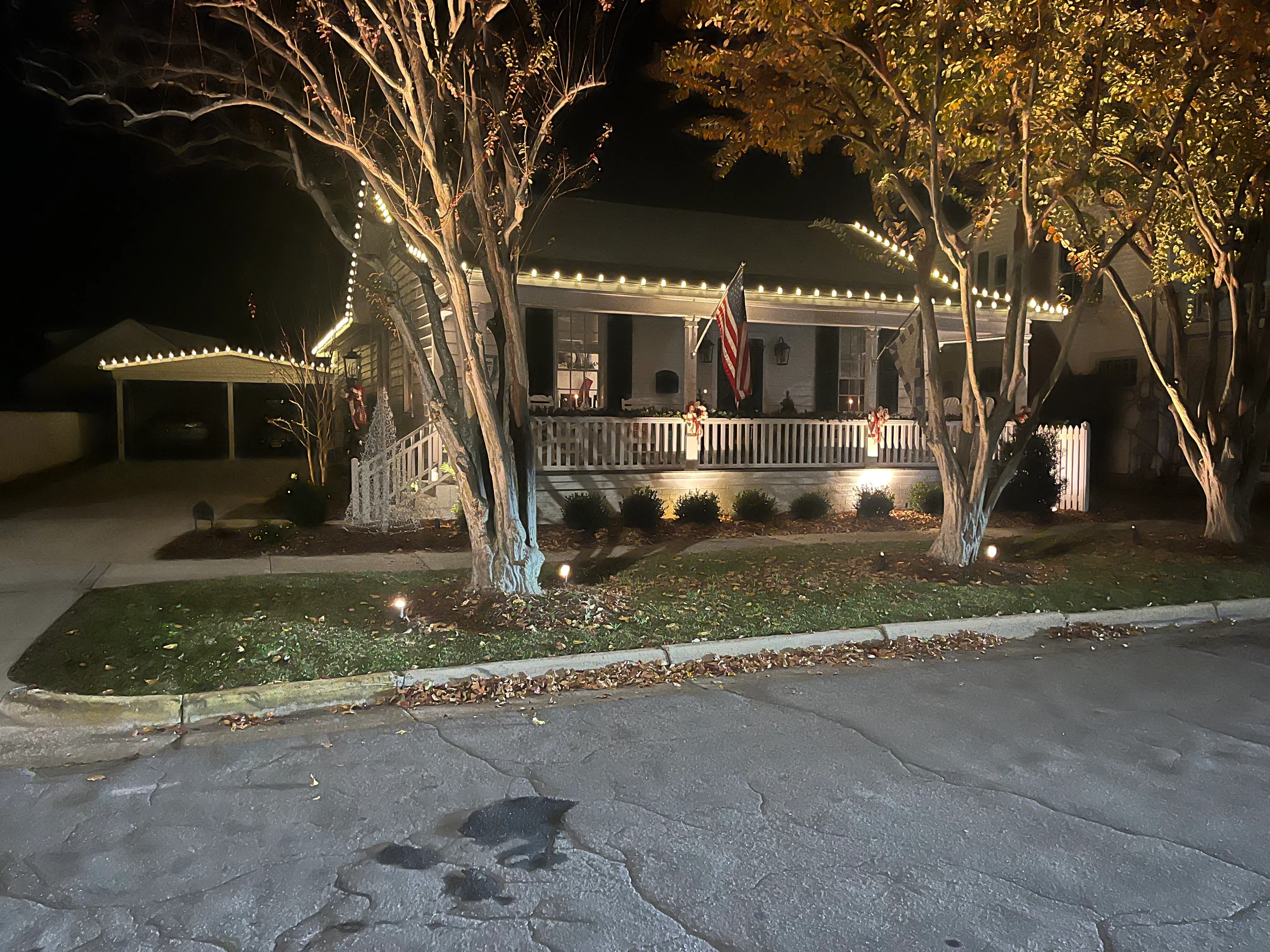 Front view of a Beaufort, NC house at night decorated with white string lights along the roofline and porch railing, illuminated trees, American flag on the porch, two candy cane decorations on the white picket fence, and a white wireframe Christmas tree near the steps installed by Walston Evans Home Services.